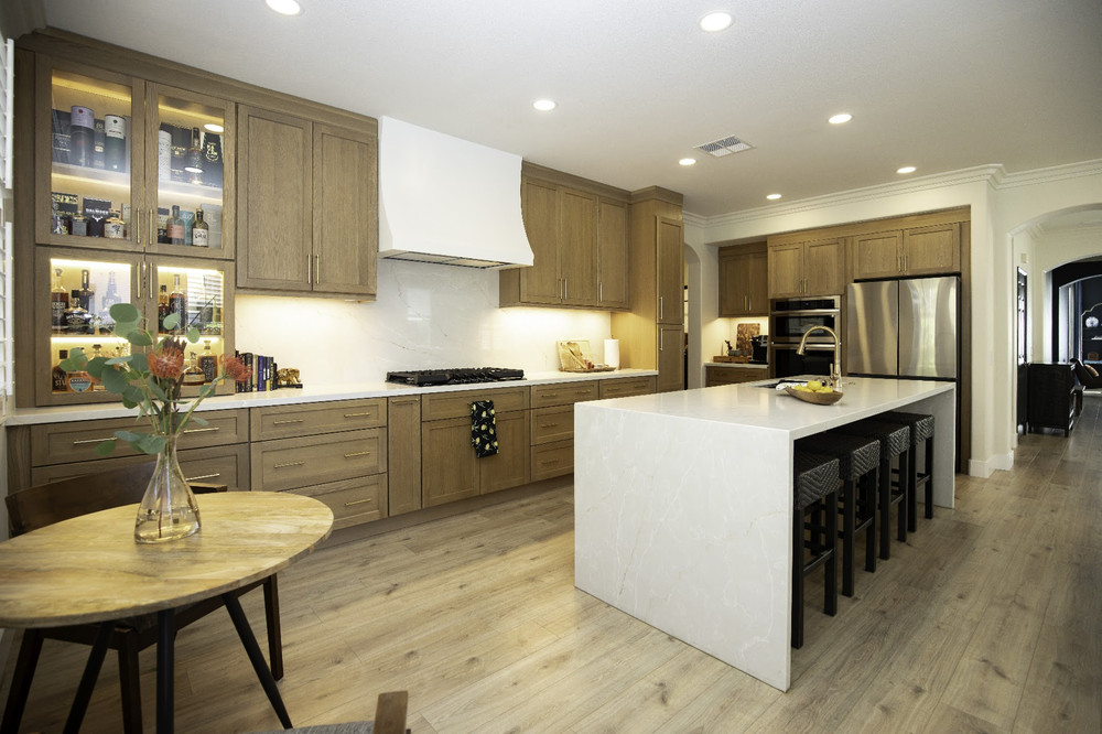 Completed kitchen with warm wood cabinetry, illuminated display cabinets, and a white waterfall island